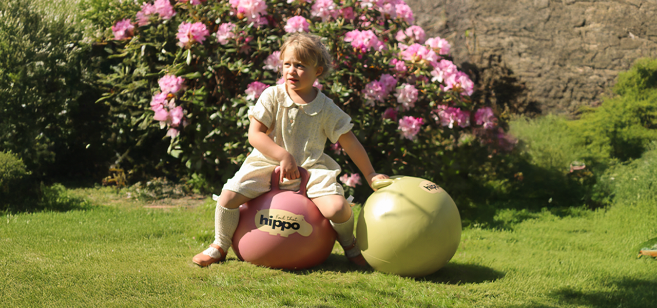 Girl sitting on two bouncy balls from Find That Hippo in a sunny garden with blooming flowers
