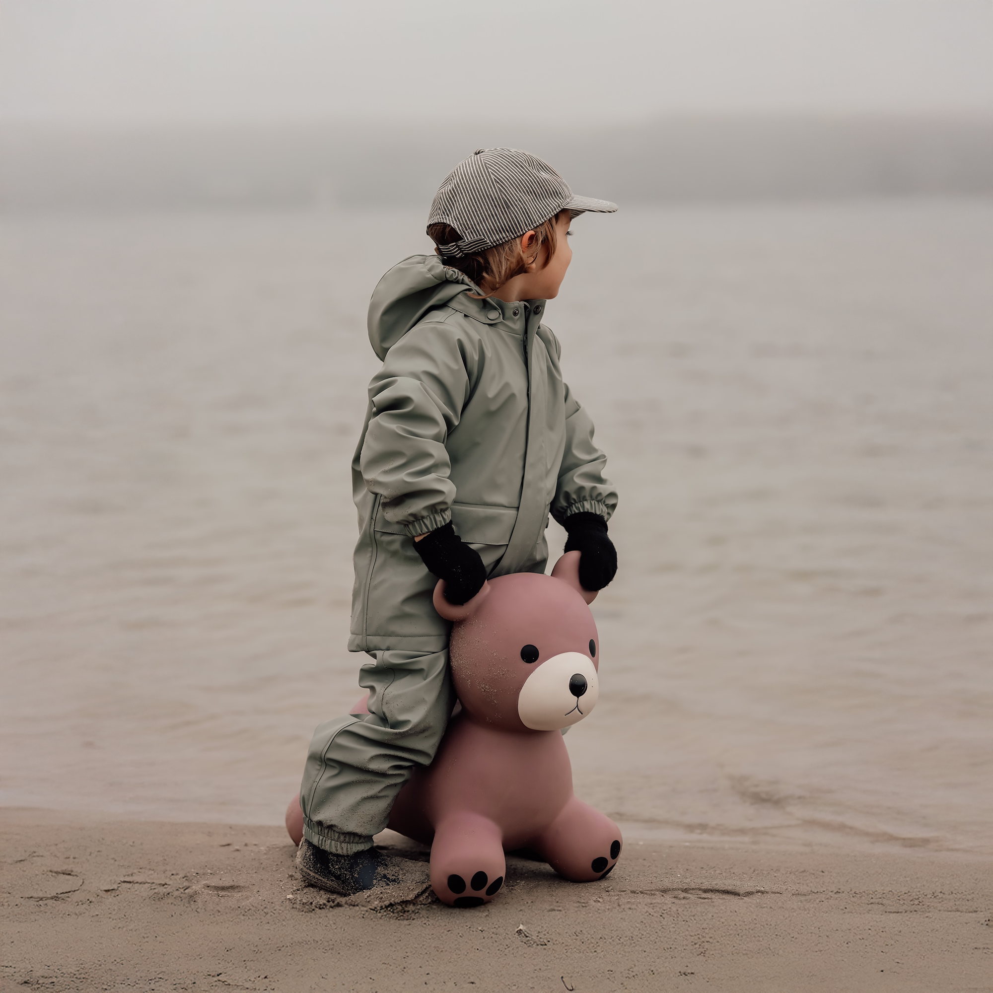 Child in green jacket riding Maya the bear bouncing toy on a sandy beach – Find That Hippo