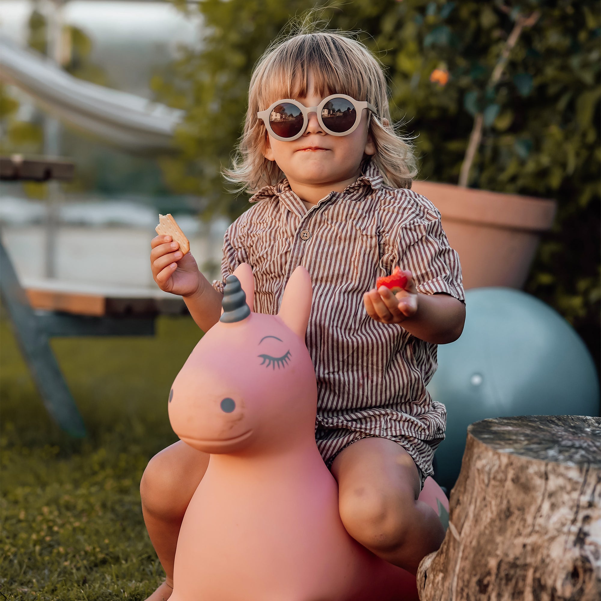 Young child wearing sunglasses sitting on Luna, the pink unicorn bouncing toy from Find That Hippo, holding snacks in a sunny garden setting