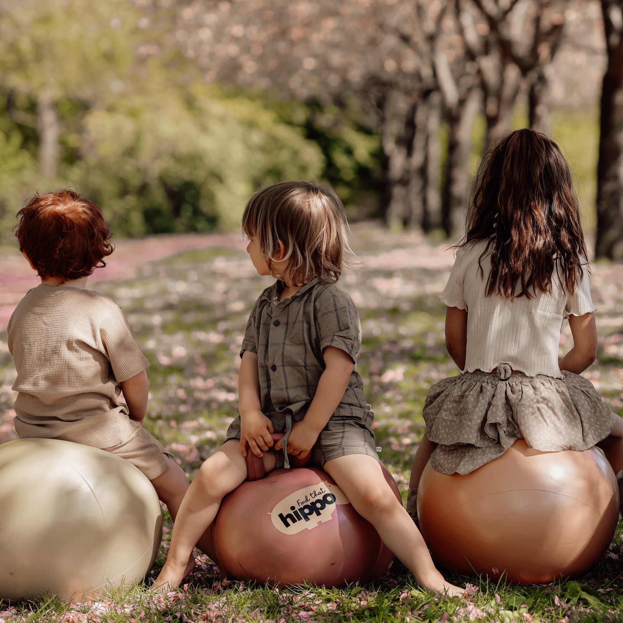 Three kids sitting on dusty pink Hippo hop balls under trees in the park – Find That Hippo