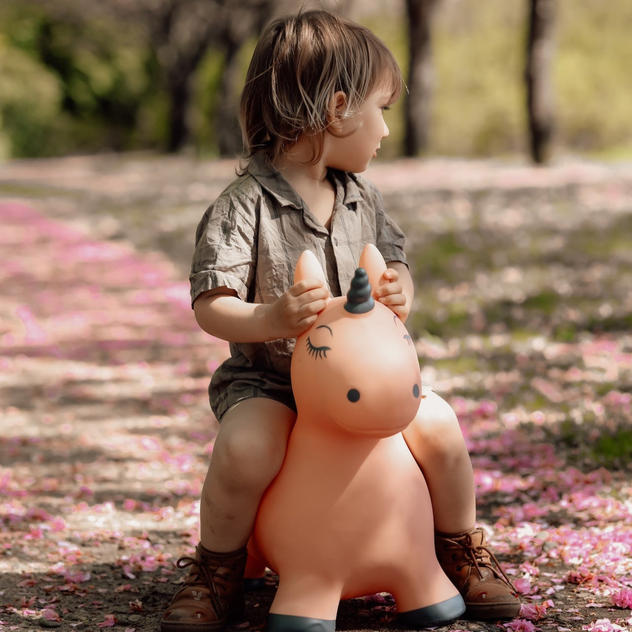 A young child sitting on Luna the Unicorn, a pink inflatable jumping toy, outdoors on a path covered in pink petals. The child is looking to the side, wearing a beige outfit and brown boots