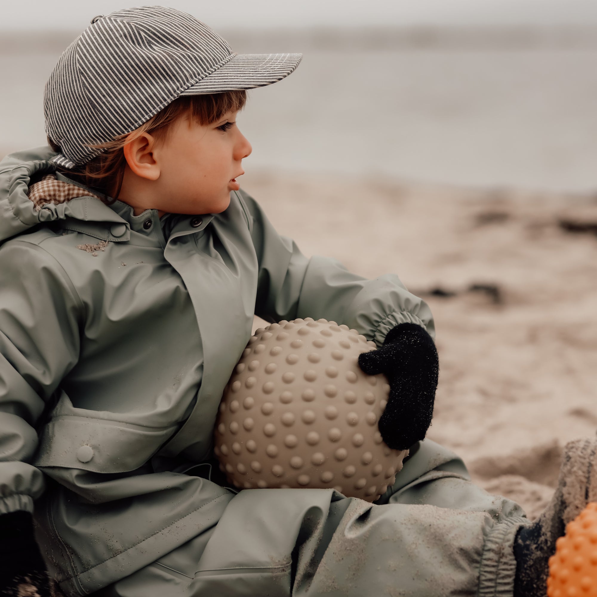 Child sitting on the beach holding a dusty green sensory ball – Find That Hippo