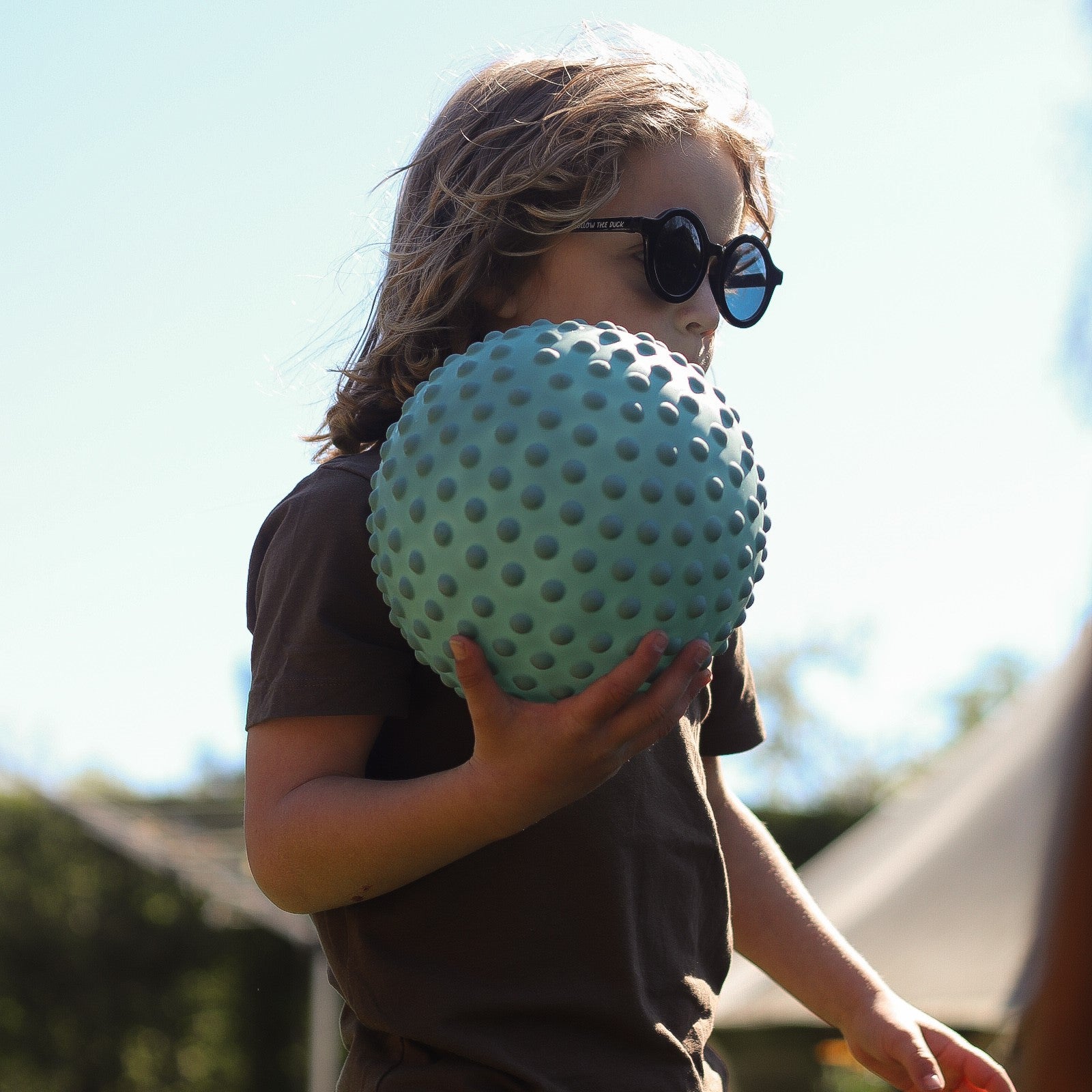 Child in sunglasses lifting a large petrol blue sensory ball outdoors in spring – Find That Hippo