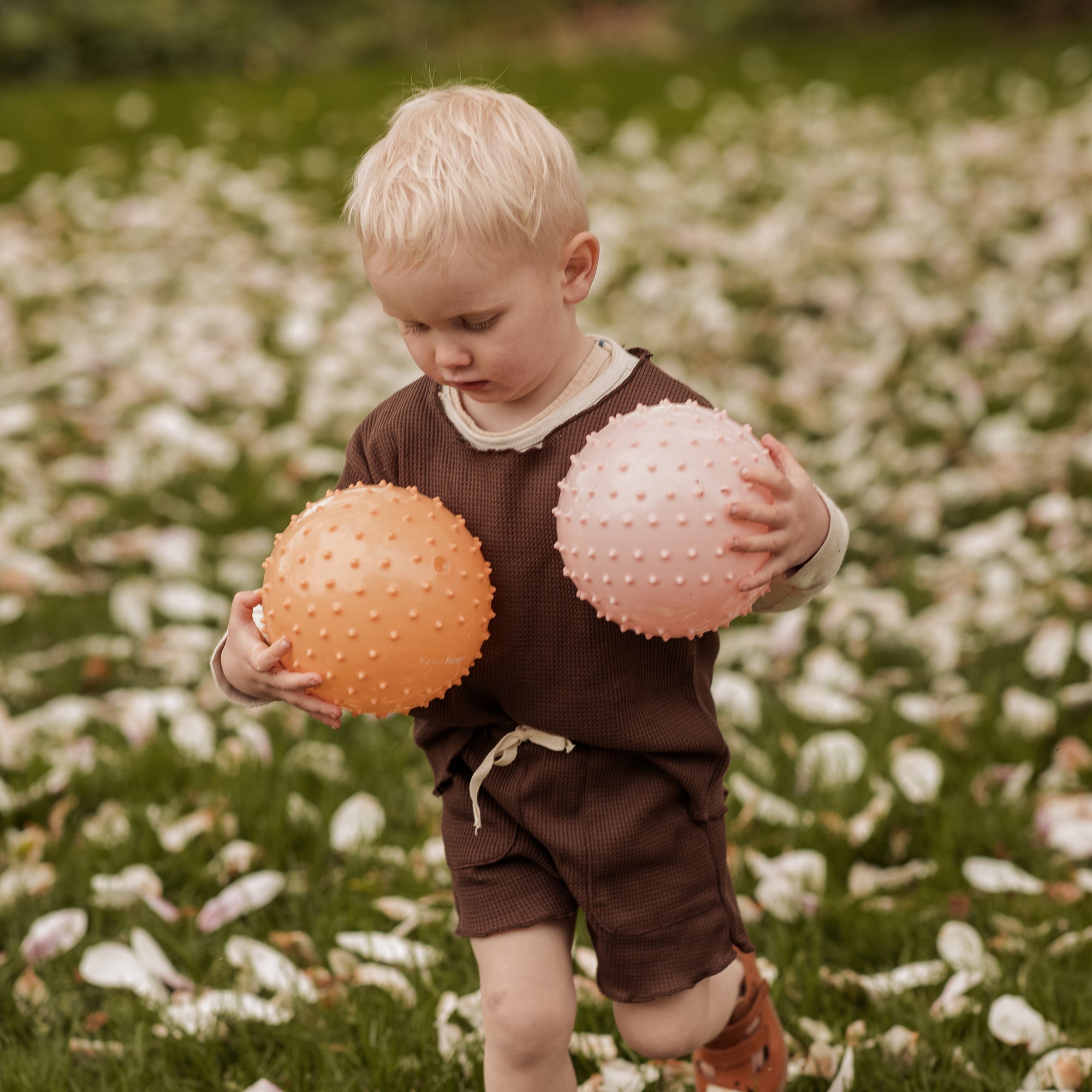 Child in brown outfit standing in a meadow, holding two sensory balls – dusty pink and muted orange – Find That Hippo