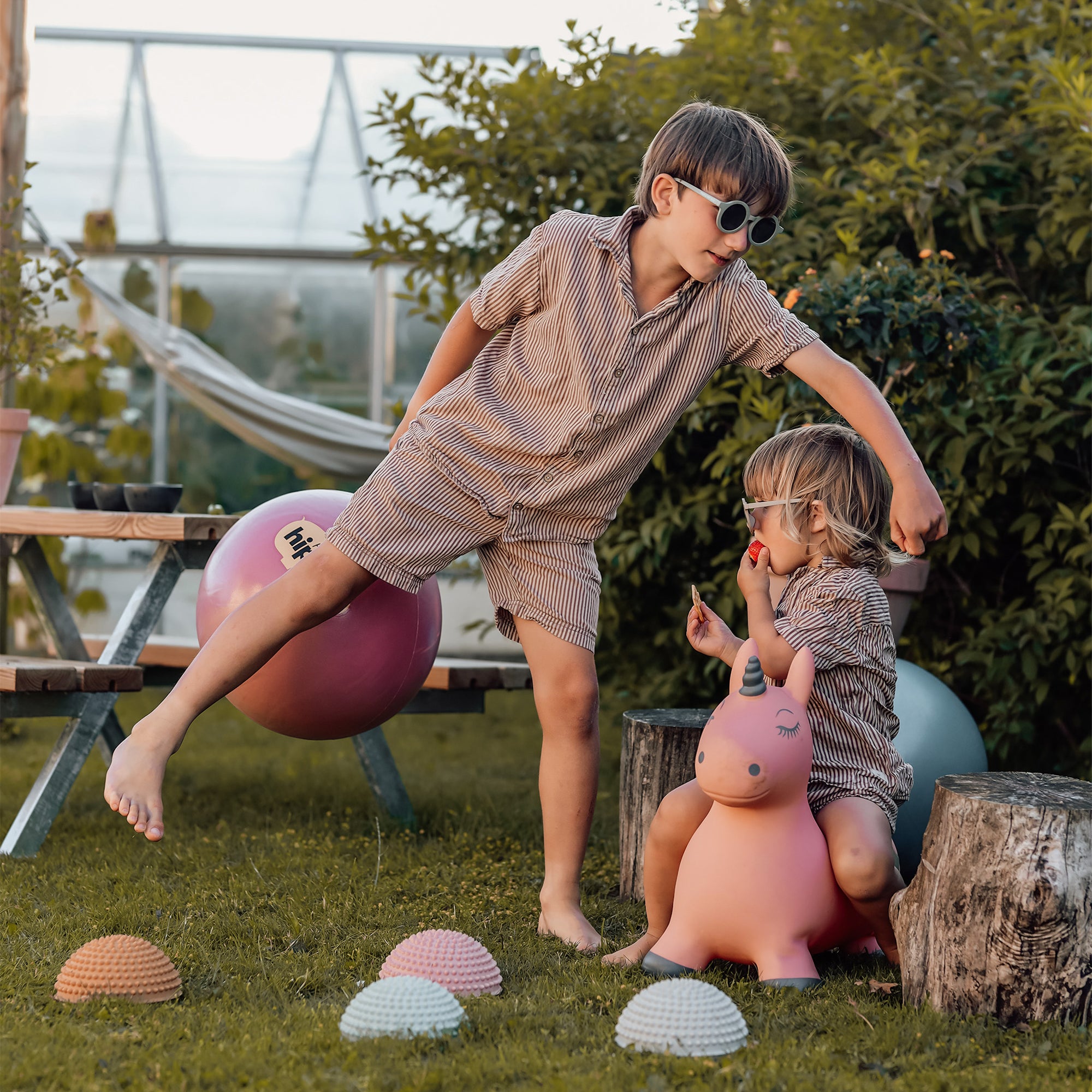 Two children playing in a garden with Luna the pink unicorn toy and pastel sensory stones from Find That Hippo; one child balances on a pink bouncy ball while the other sits on Luna eating snacks